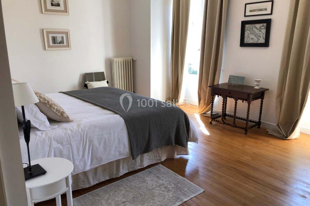 Chambre lumineuse avec lit double, parquet en bois, table ancienne et décorations murales encadrées.