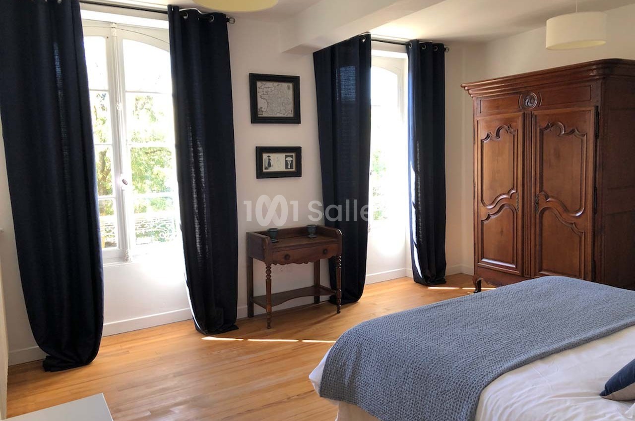 Chambre lumineuse avec parquet, armoire en bois, lit avec couverture bleue et rideaux sombres encadrant deux fenêtres.