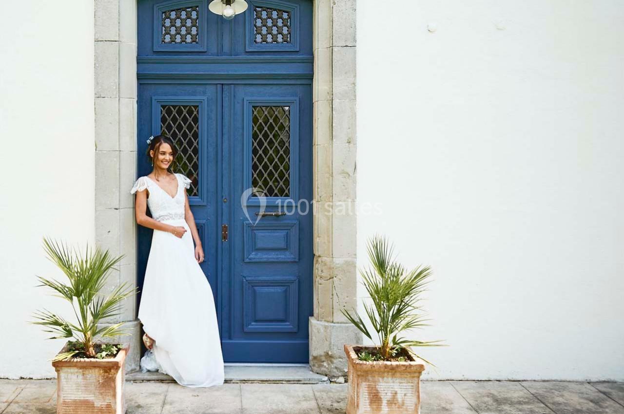 Femme en robe blanche debout devant une porte bleue encadrée de plantes en pot sur une terrasse ensoleillée.
