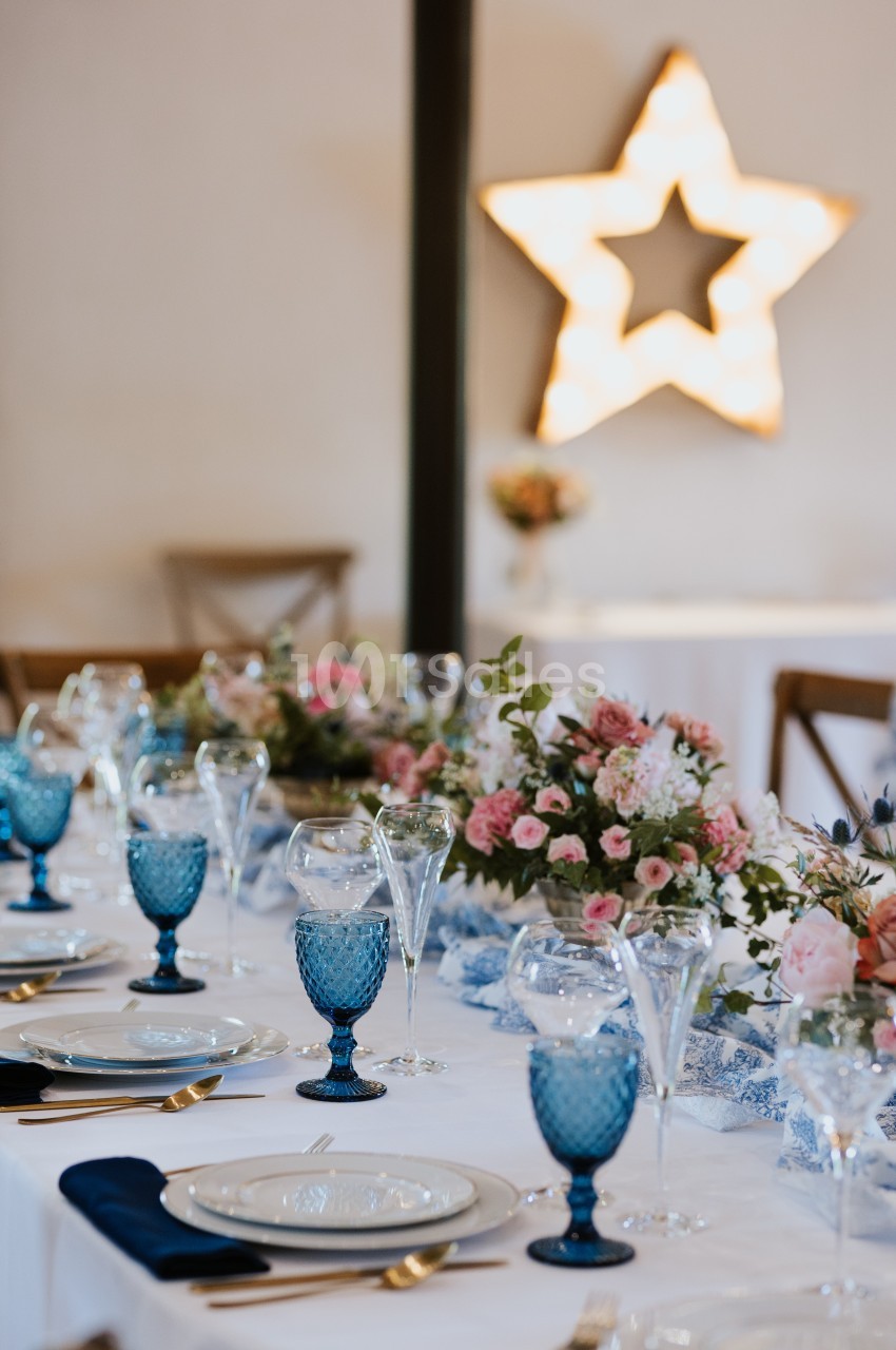Table élégamment dressée avec vaisselle bleue, bouquets de fleurs roses et étoile lumineuse en arrière-plan.