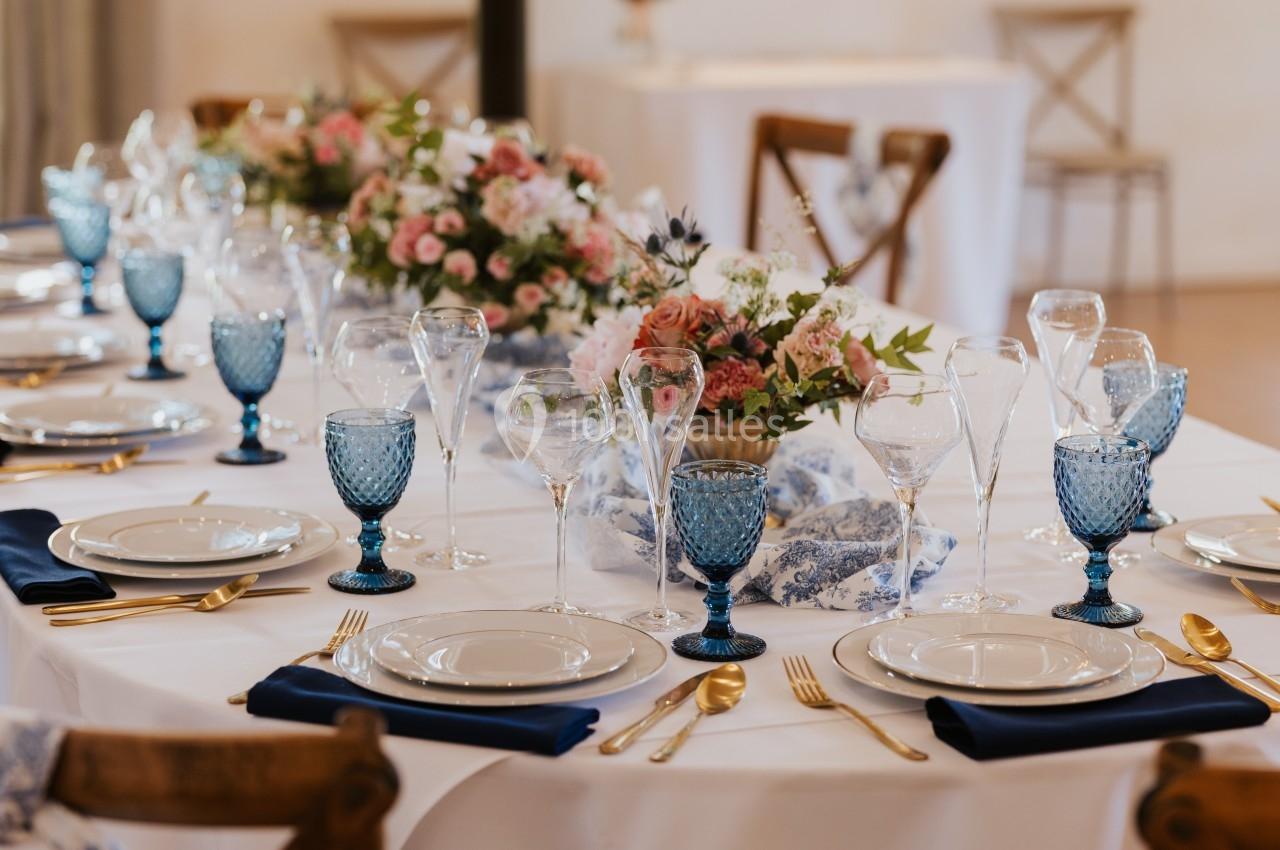 Table élégamment dressée avec vaisselle blanche, verres bleus, couverts dorés et centre de table floral.