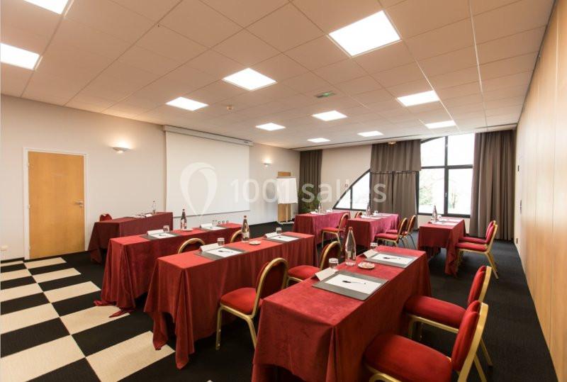 Salle de réunion lumineuse avec tables rouges alignées, chaises assorties, écran de projection et tableau blanc.
