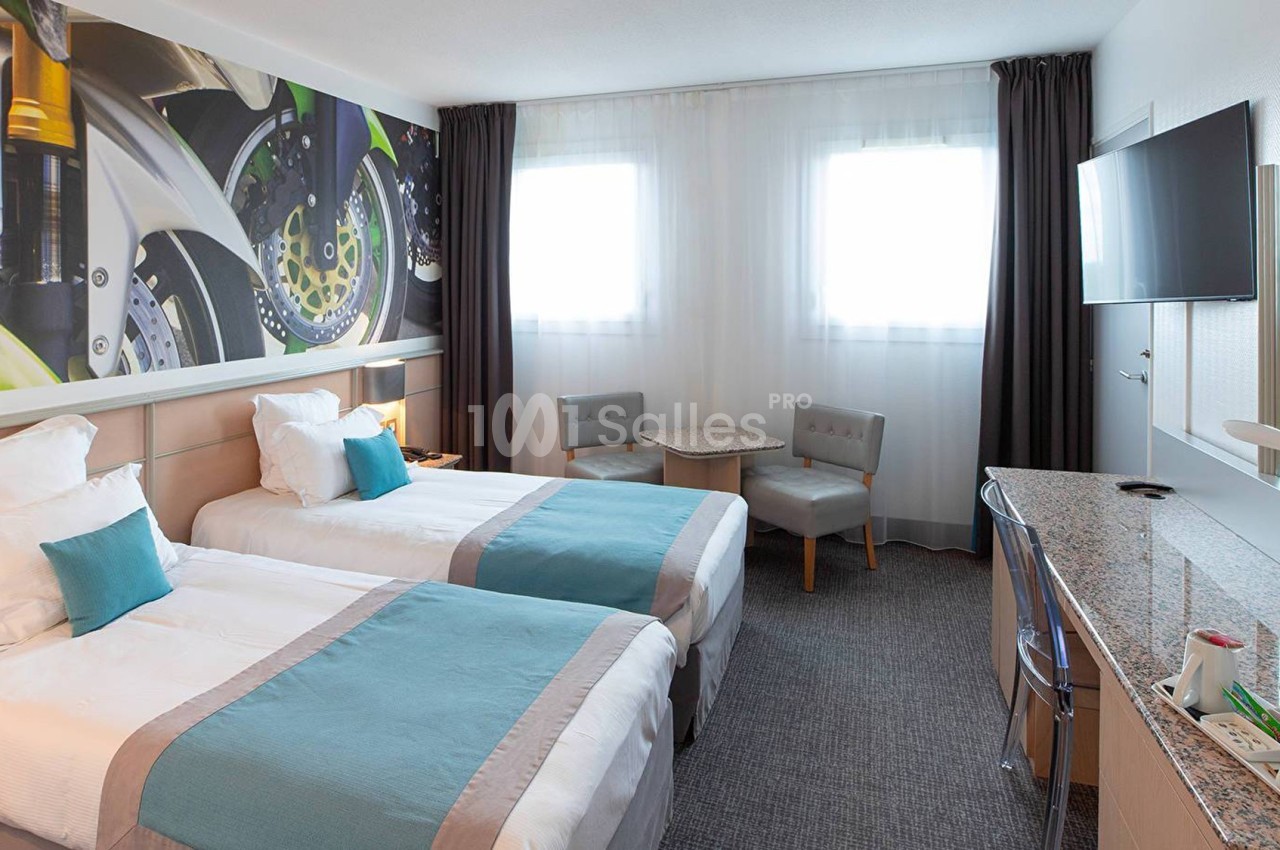 Chambre d'hôtel lumineuse avec deux lits simples, un bureau, une télévision murale et une décoration murale colorée.