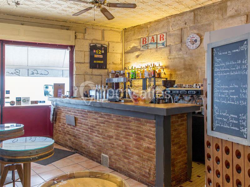 Location salle Bordeaux (Gironde) - Restaurant Le Family #9 Intérieur d'un café avec un comptoir en briques, des bouteilles alignées, un tableau de menu et des tables rondes en bois.