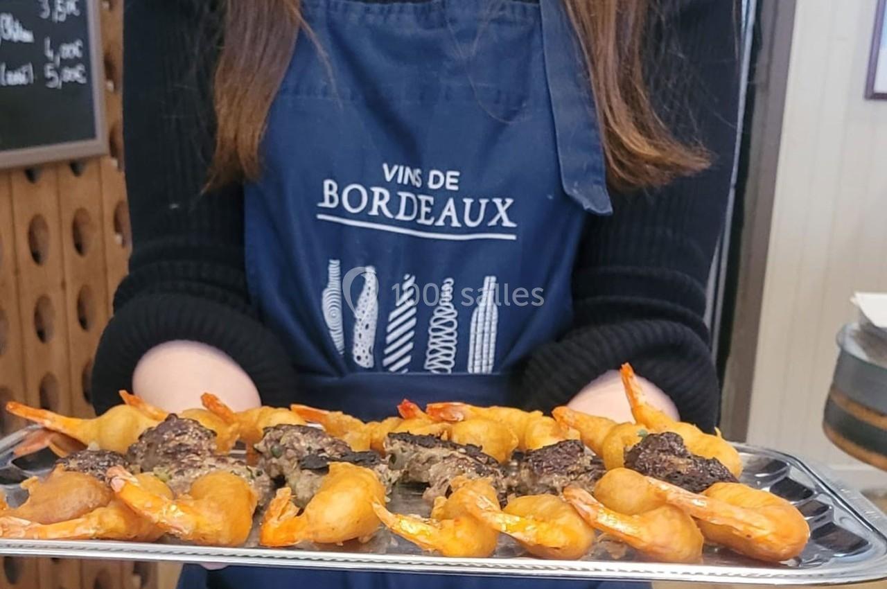Location salle Bordeaux (Gironde) - Restaurant Le Family #7 Plateau de bouchées apéritives avec crevettes et viande, tenu par une personne portant un tablier ’Vins de Bordeaux’.