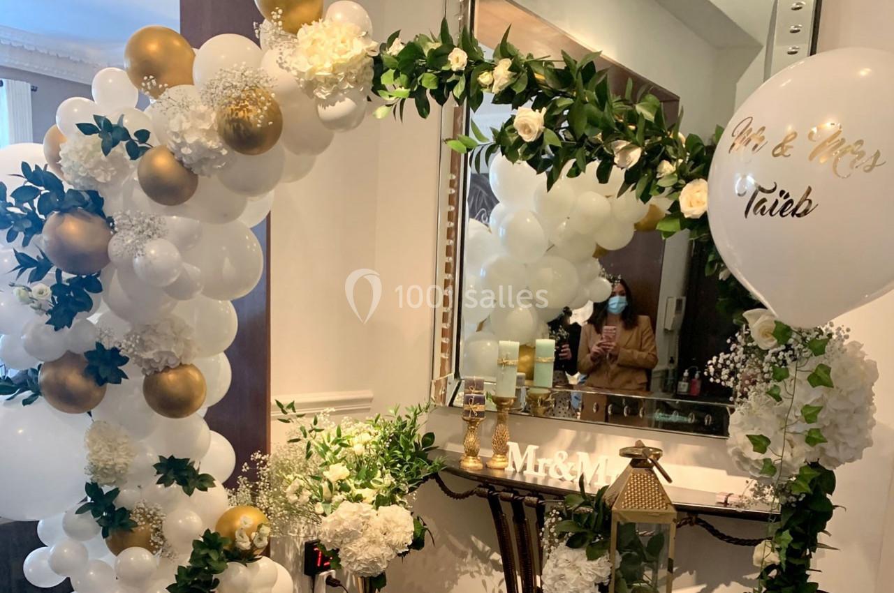 Décoration de mariage avec arche de ballons blancs et dorés, fleurs blanches, et ballon personnalisé près d'un miroir.