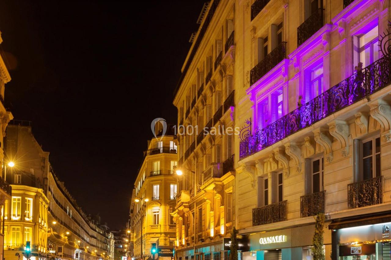 Rue animée de nuit avec des bâtiments illuminés, dont une façade éclairée en violet, et des boutiques au rez-de-chaussée.