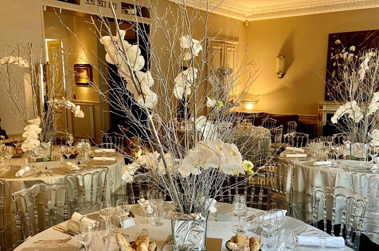 Salle de réception élégante avec tables dressées, décorations florales blanches et chaises transparentes.