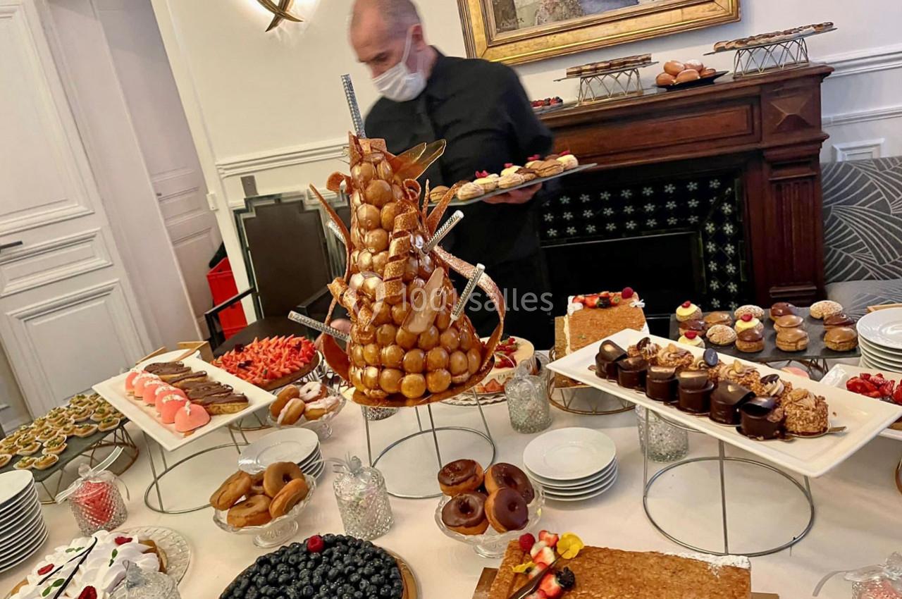 Table garnie de pâtisseries variées, avec un serveur masqué en arrière-plan près d'une cheminée décorative.