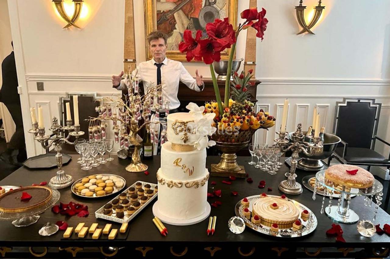 Table décorée avec des desserts variés, un gâteau blanc central, des fleurs rouges et un homme en arrière-plan.
