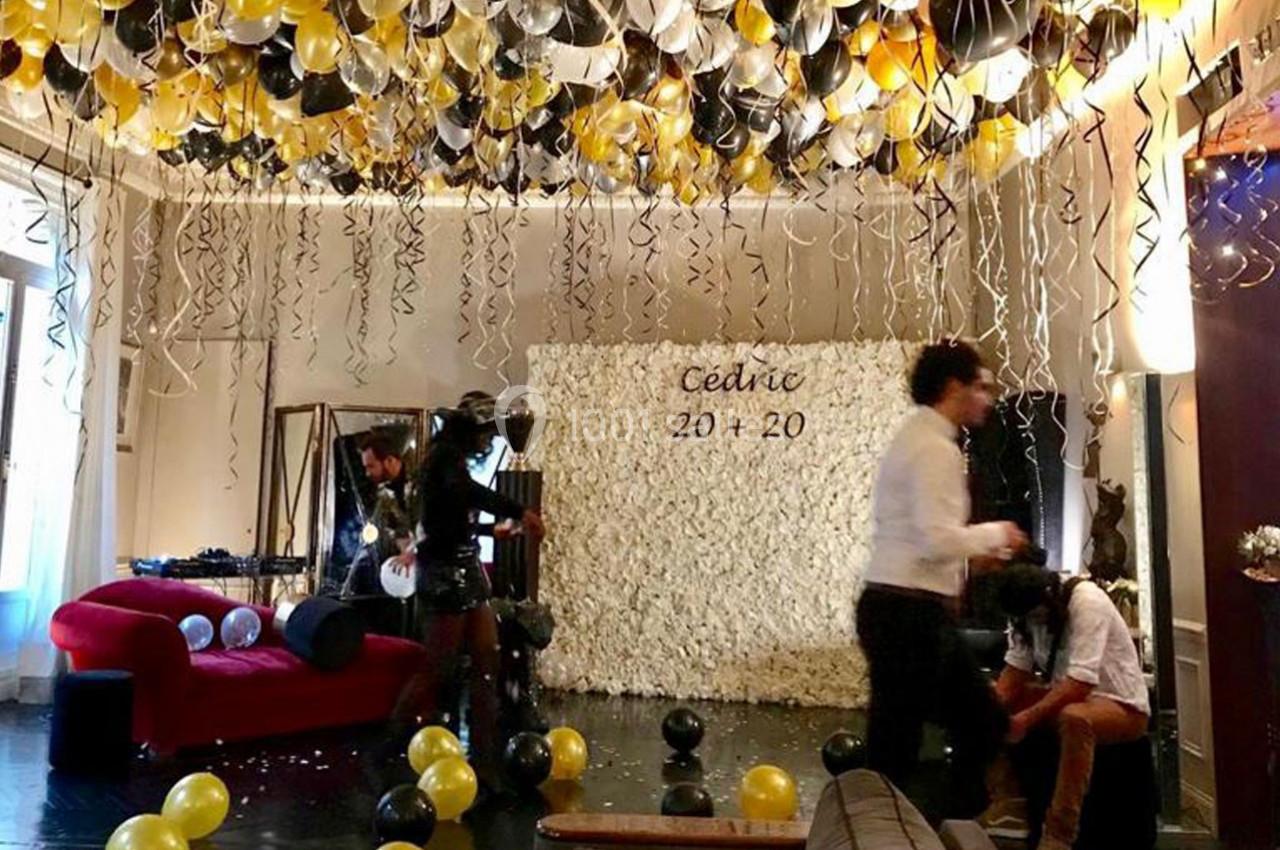 Salle décorée pour une fête avec ballons noirs, dorés et blancs, mur de fleurs blanches et inscription ’Cédric 20 + 20’.