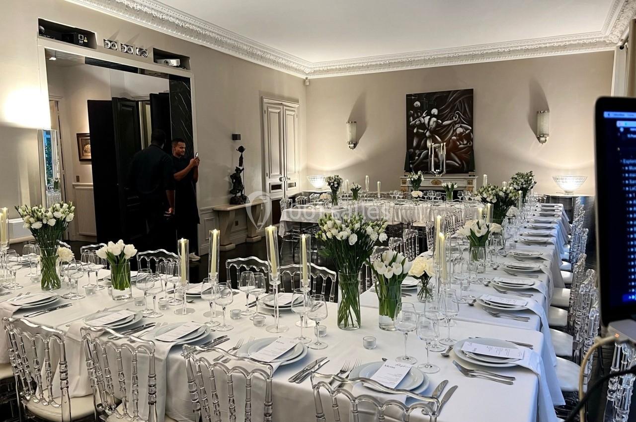 Salle élégante avec deux longues tables dressées, décorées de fleurs blanches et de bougies, prête pour un événement.
