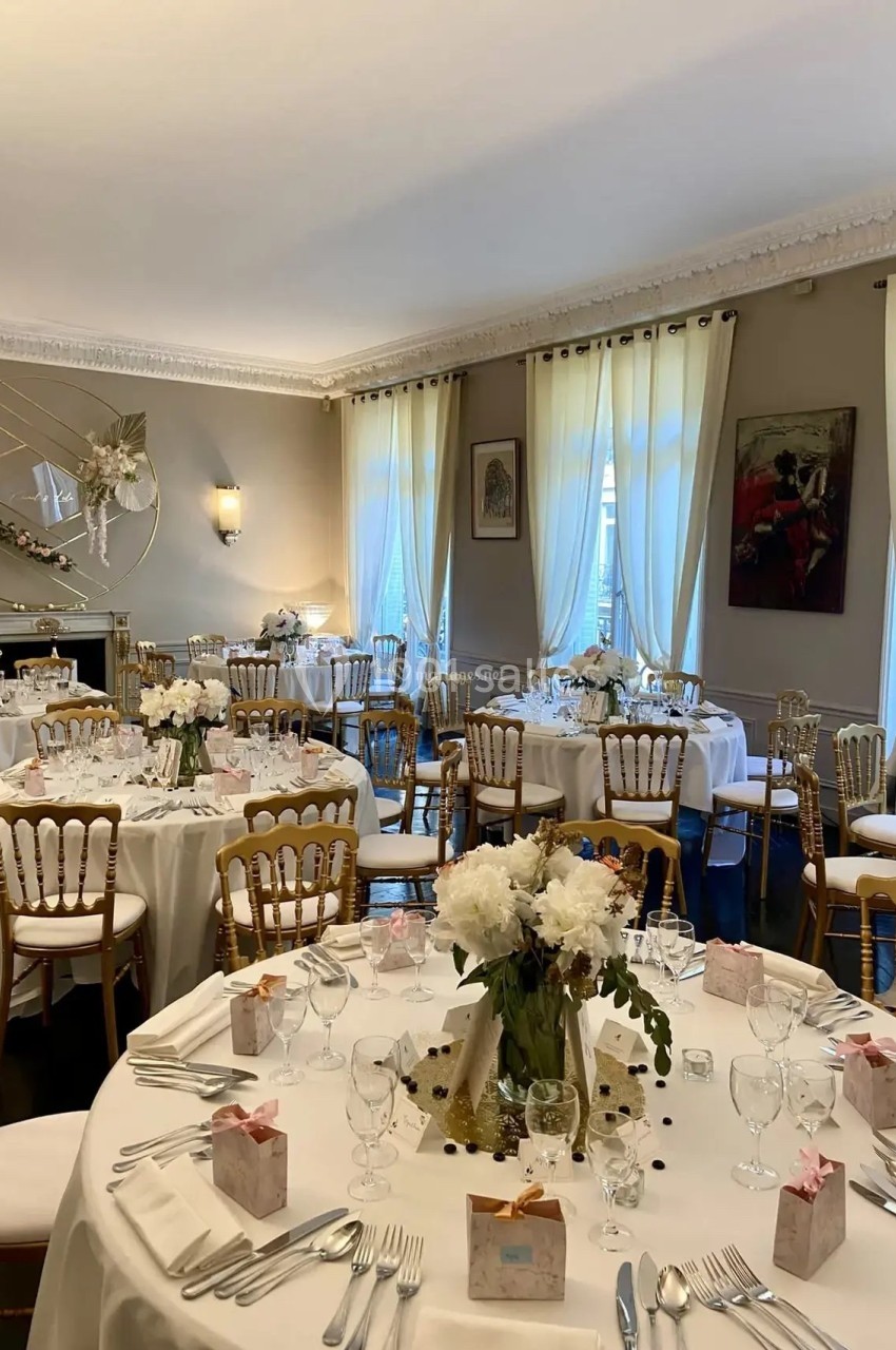 Salle de réception élégamment décorée avec des tables rondes, nappes blanches, chaises dorées et arrangements floraux.