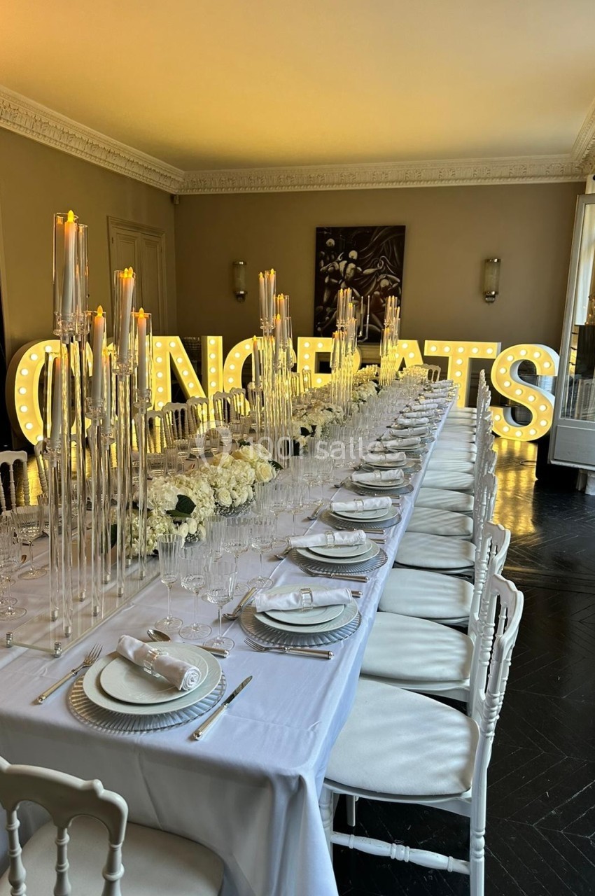 Table élégamment dressée avec chandeliers et fleurs blanches, devant des lettres lumineuses formant ’CONGRATS’.