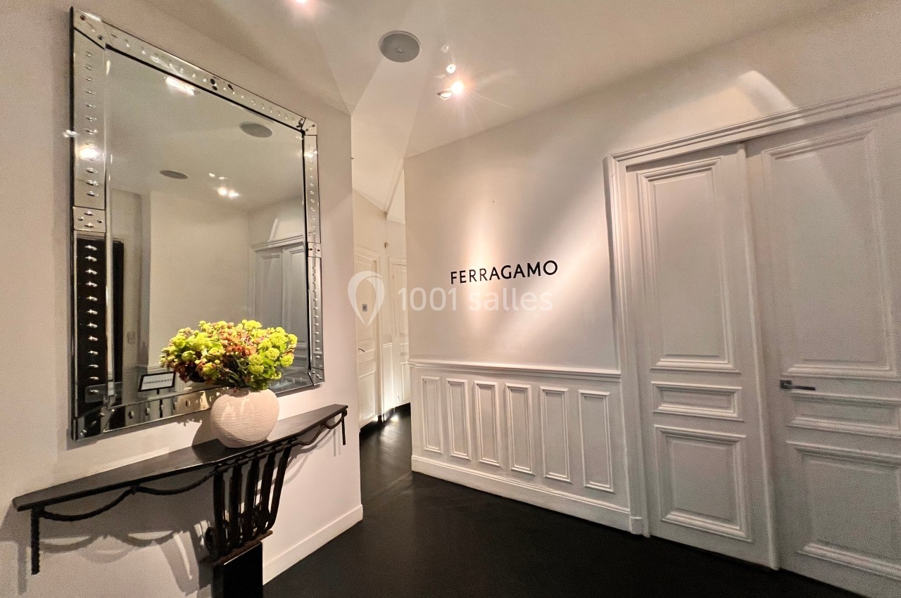Entrée élégante avec mur blanc, miroir encadré, console noire, vase de fleurs et logo ’Ferragamo’ sur le mur.