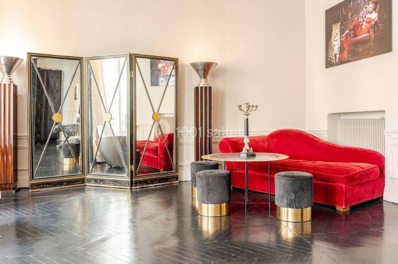 Salon élégant avec canapé rouge, miroirs décoratifs, tabourets gris et sol en parquet sombre.