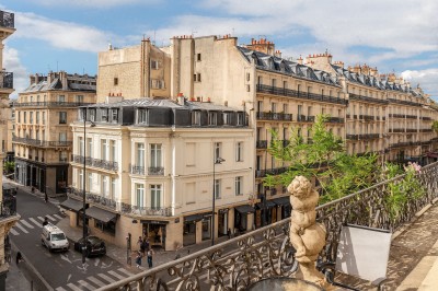 Une statue en pierre orne un balcon avec vue sur des immeubles anciens dans un environnement urbain.