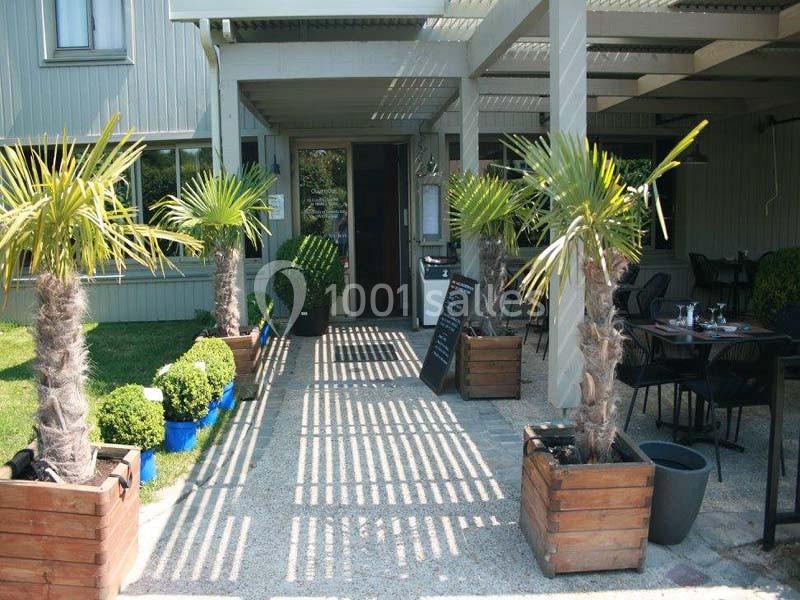 Entrée d'un restaurant avec terrasse ombragée, palmiers en pots et tables dressées visibles sur le côté.
