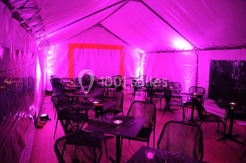 Intérieur d'une tente éclairée par des lumières roses, avec des tables et des chaises disposées pour un événement.