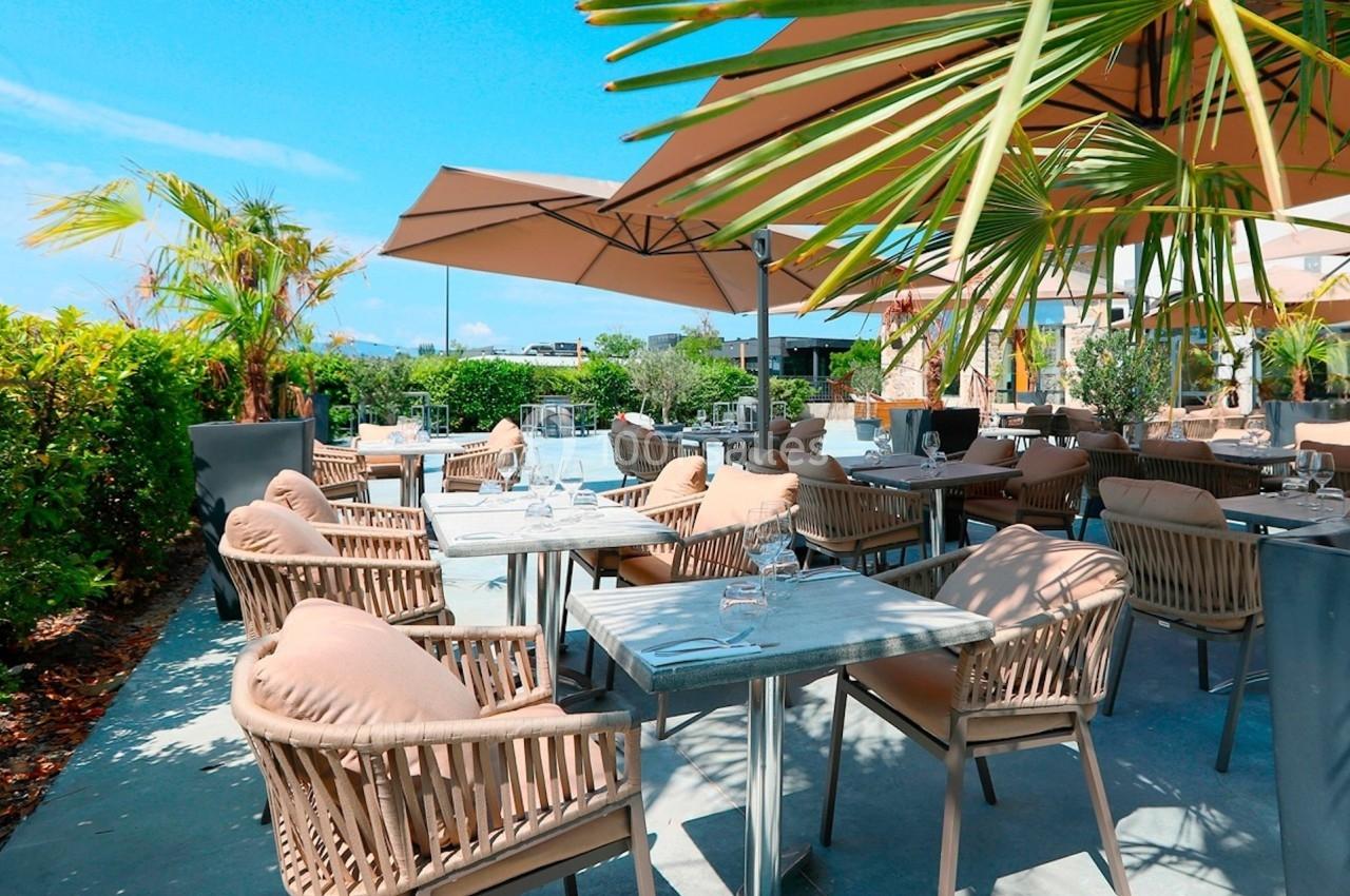 Terrasse extérieure d'un restaurant avec tables, chaises en rotin, parasols et végétation sous un ciel bleu.