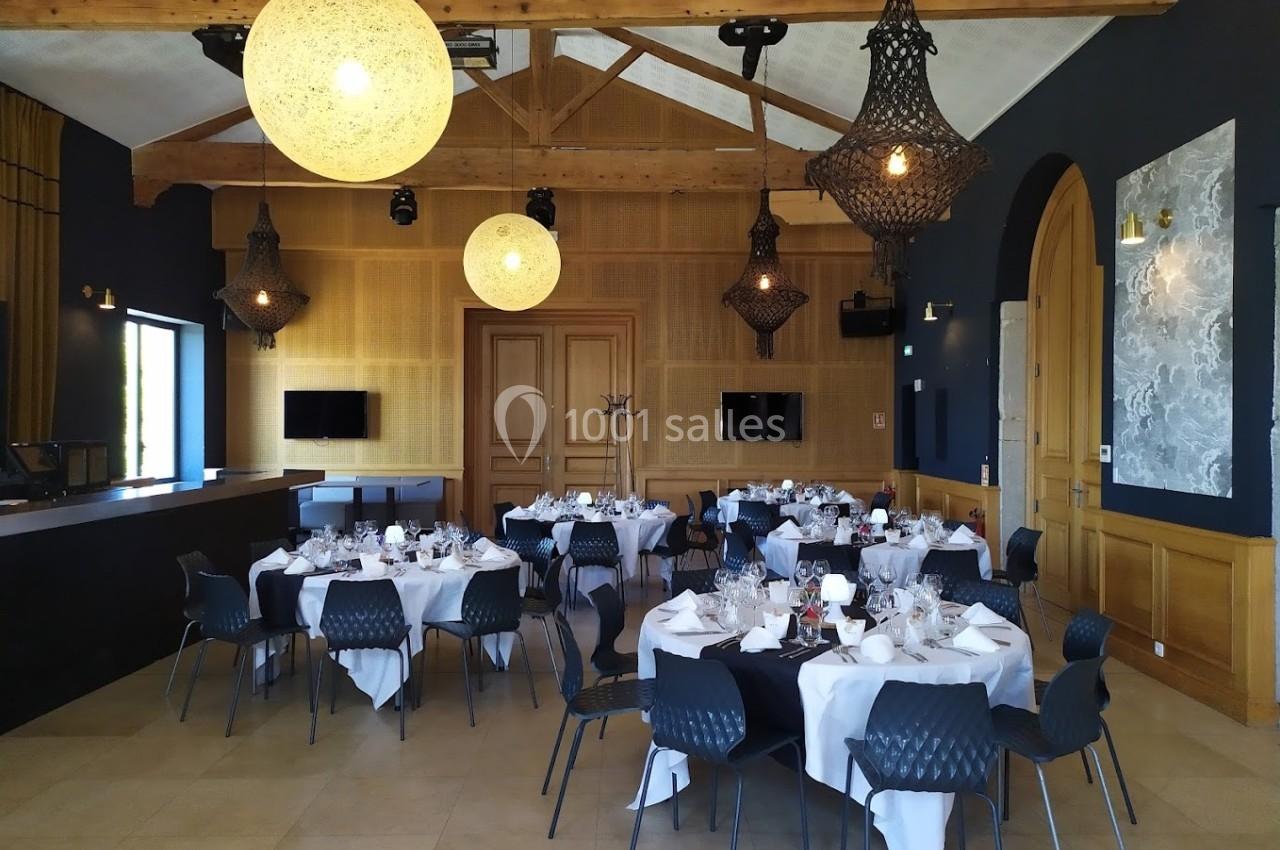 Salle de réception élégante avec tables rondes dressées, lustres suspendus et poutres apparentes en bois.