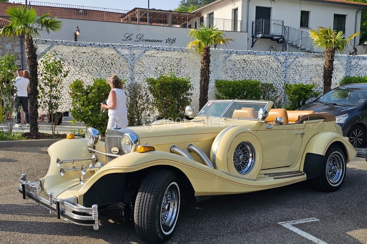 Voiture ancienne beige décapotable stationnée devant un bâtiment avec des palmiers et des personnes en arrière-plan.