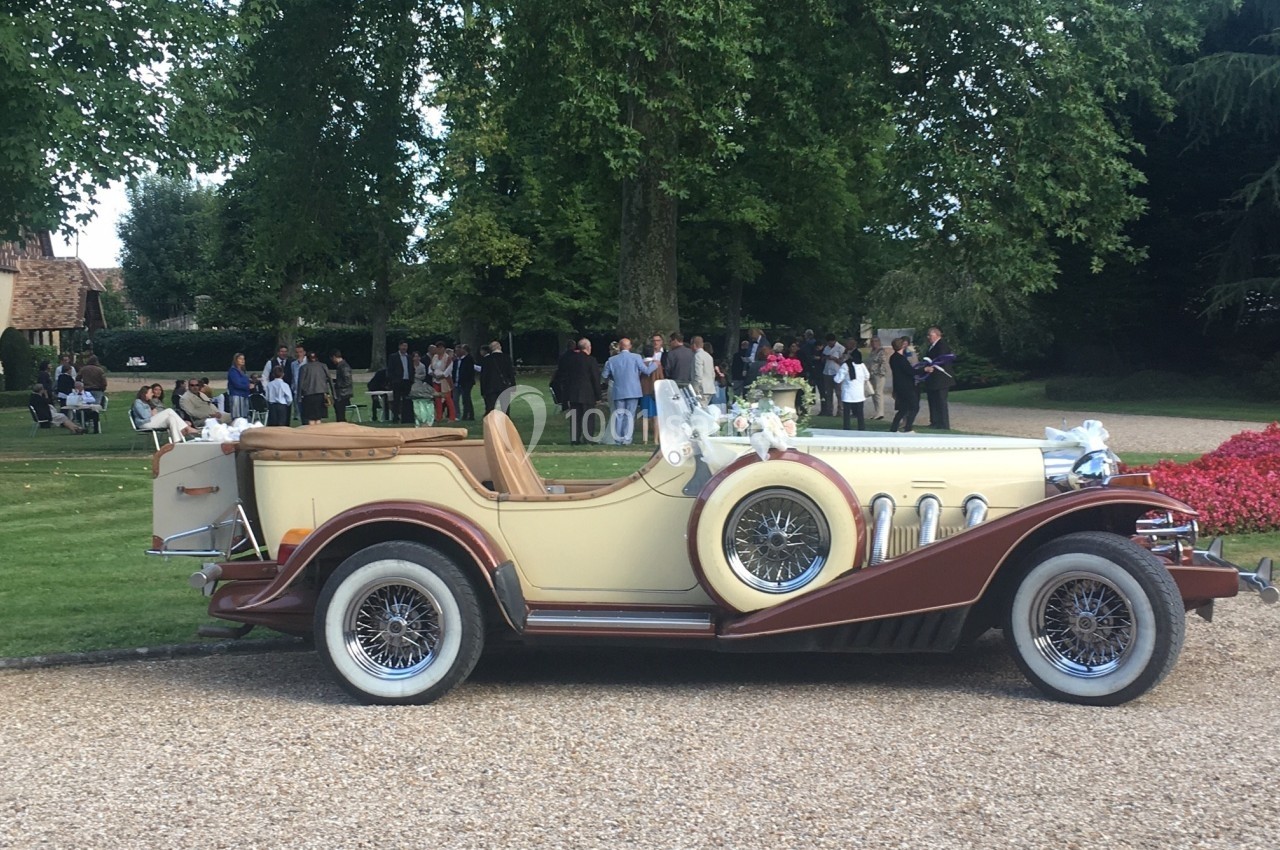 Voiture ancienne beige et marron décorée de fleurs blanches, garée sur une allée gravillonnée devant un parc avec des…