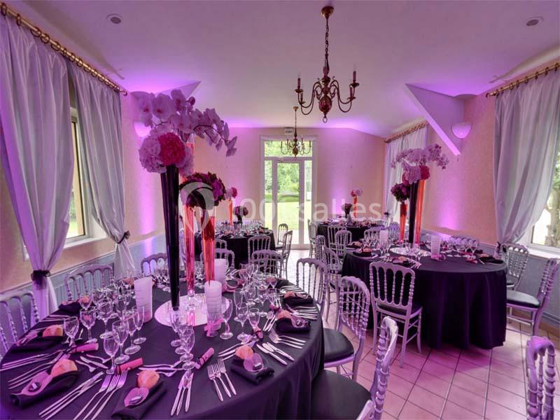 Salle de réception décorée avec des tables rondes, nappes noires, fleurs roses et éclairage d'ambiance violet.