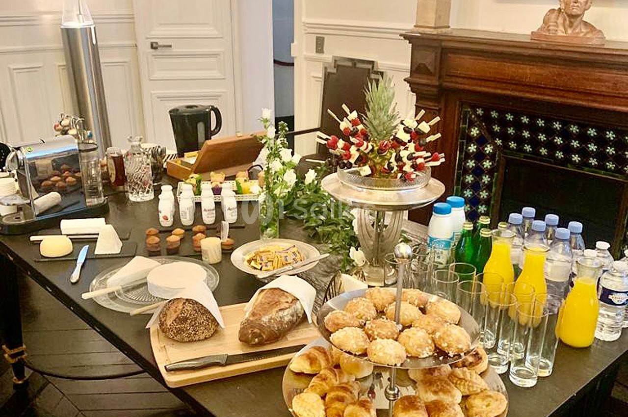 Table garnie de viennoiseries, pains, jus, boissons et fruits, disposée dans une salle élégante avec cheminée en arrière…