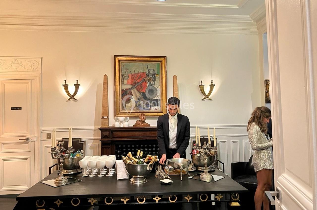 Un homme en costume prépare une table avec des boissons et des coupes dans une pièce élégante et bien éclairée.