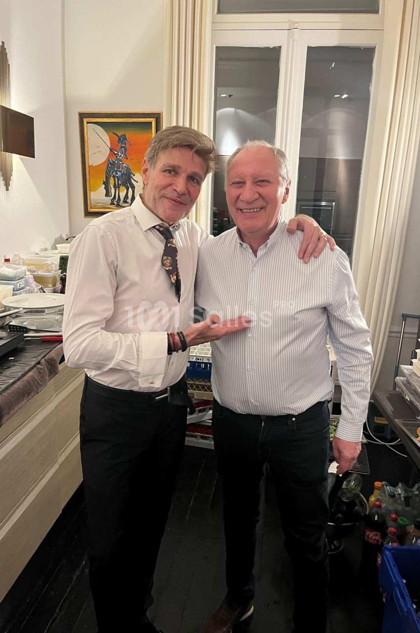 Deux hommes souriants posent côte à côte dans une pièce avec des tables remplies de plats et de boissons.