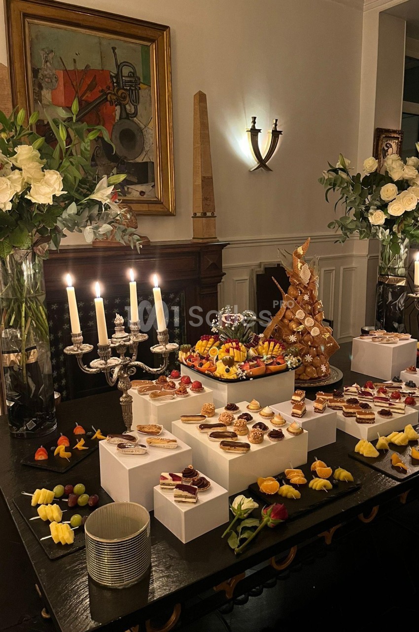 Table garnie de desserts variés, pièces montées et bougies allumées, entourée de bouquets de fleurs blanches.