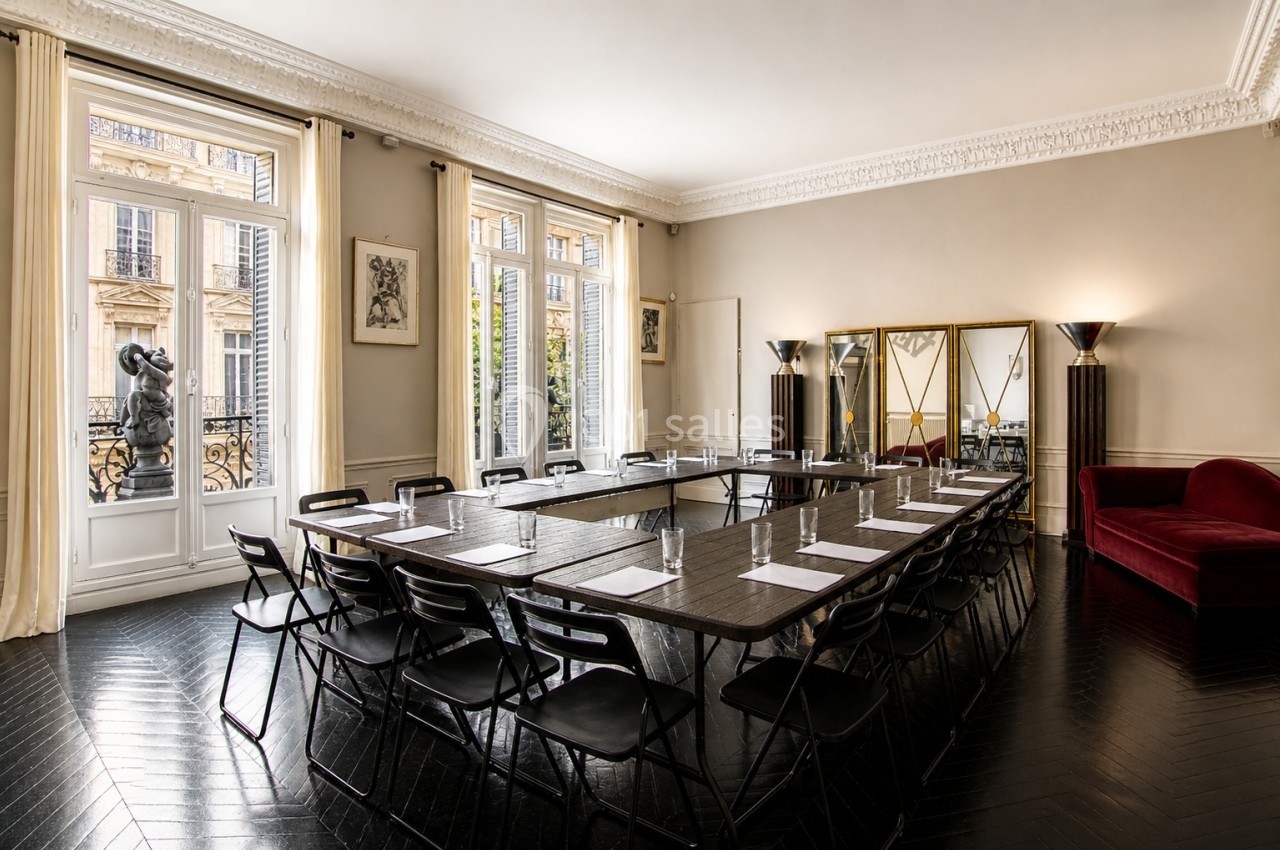 Salle de réunion lumineuse avec table en U, chaises noires, grandes fenêtres et miroirs sur un mur beige.