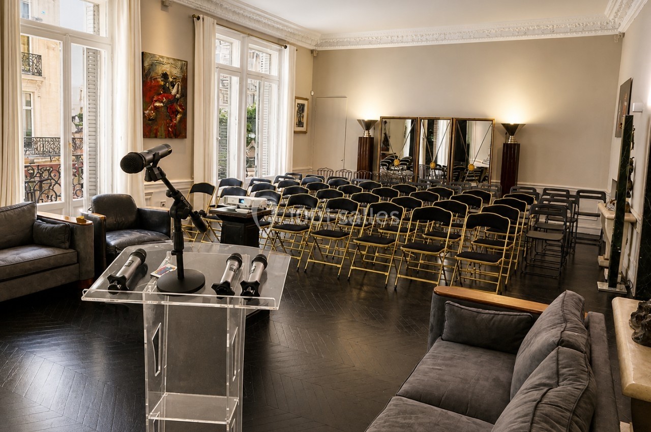 Salle lumineuse avec chaises alignées, pupitre transparent équipé de micros, canapés et grandes fenêtres donnant sur l…