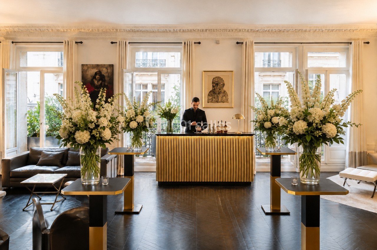 Un barman prépare des boissons derrière un comptoir doré dans un salon élégant décoré de grandes compositions florales.