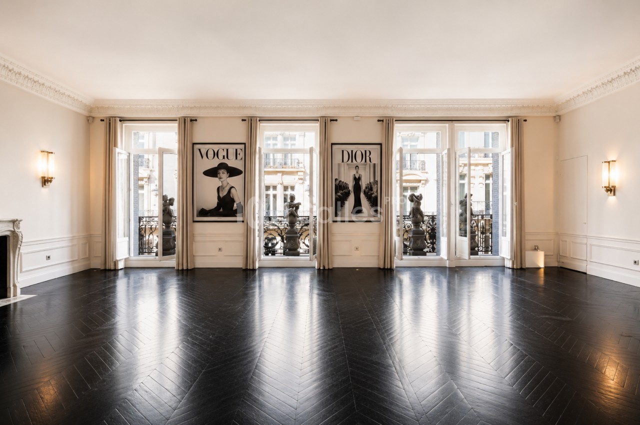 Salle lumineuse avec parquet en chevrons, grandes fenêtres donnant sur un balcon et affiches décoratives au mur.
