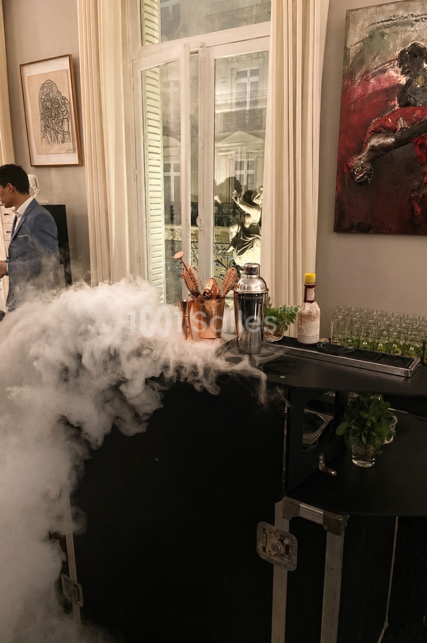 Bar avec fumée, contenant en cuivre rempli de bouteilles, shakers, herbes fraîches et verrerie devant une fenêtre.