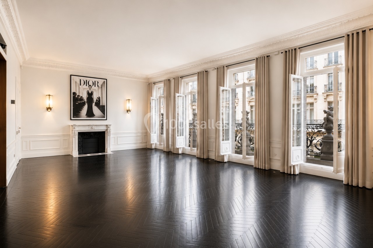 Salon spacieux avec parquet sombre, cheminée, grandes fenêtres ouvertes et vue sur des immeubles haussmanniens.