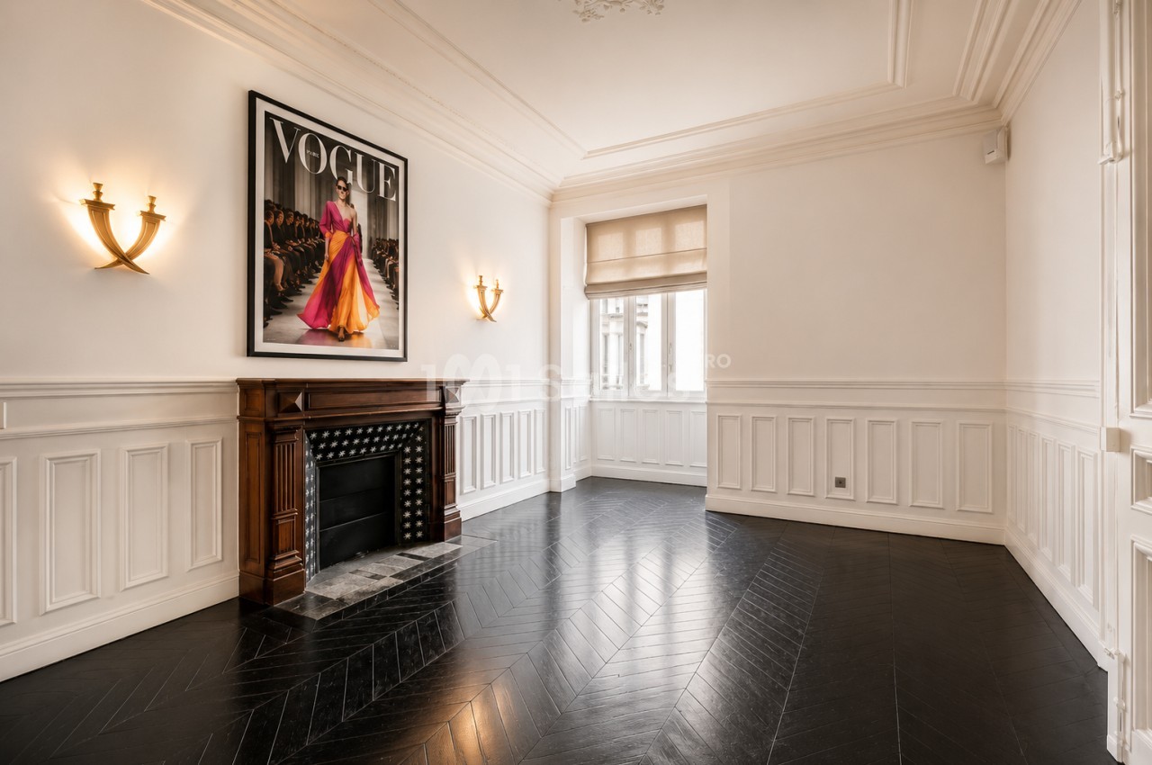 Salle lumineuse avec parquet en chevrons, cheminée encadrée de deux appliques et grande affiche murale.