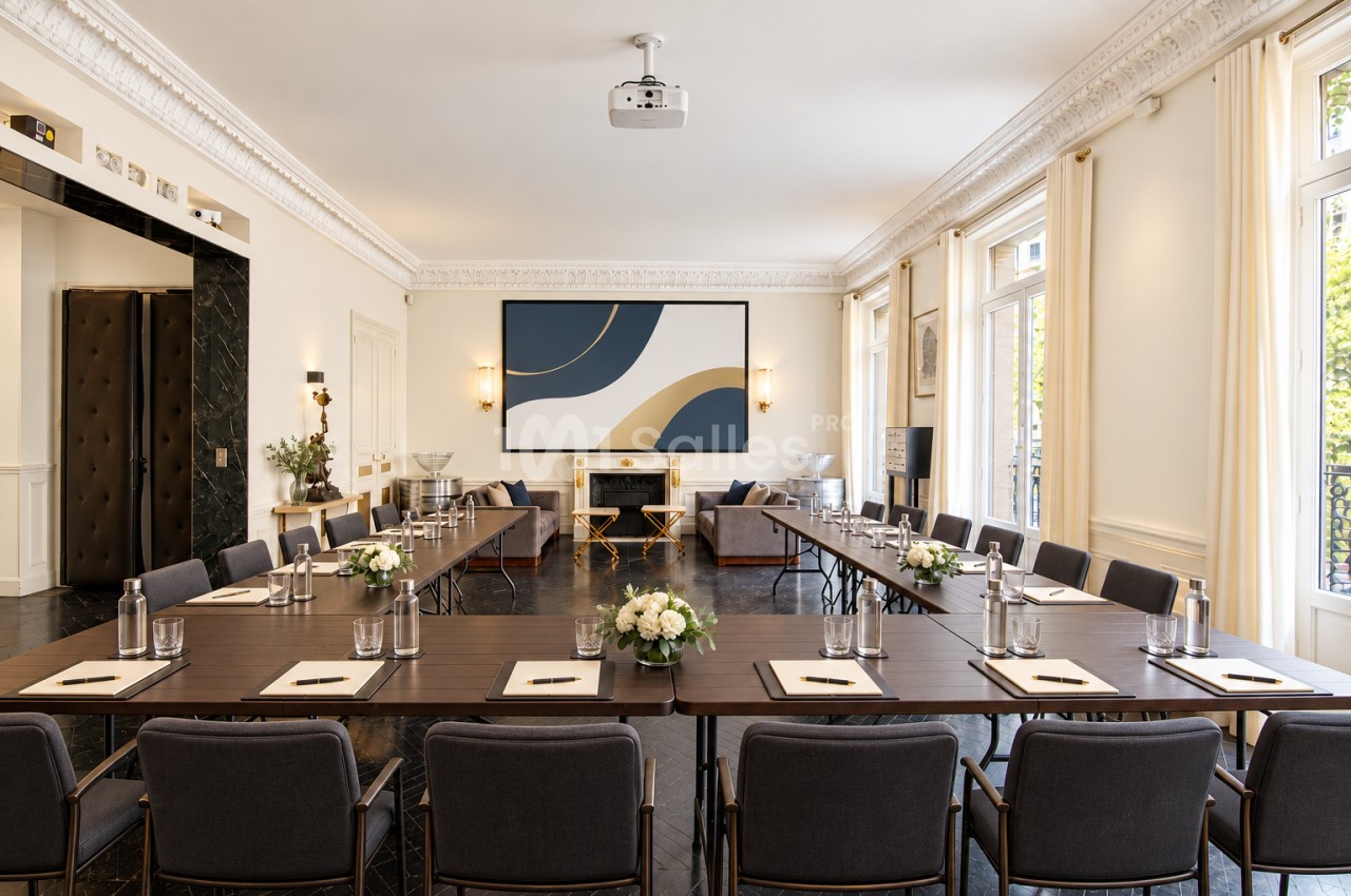 Salle de réunion lumineuse avec tables en U, chaises grises, décor élégant et grandes fenêtres laissant entrer la lumière…