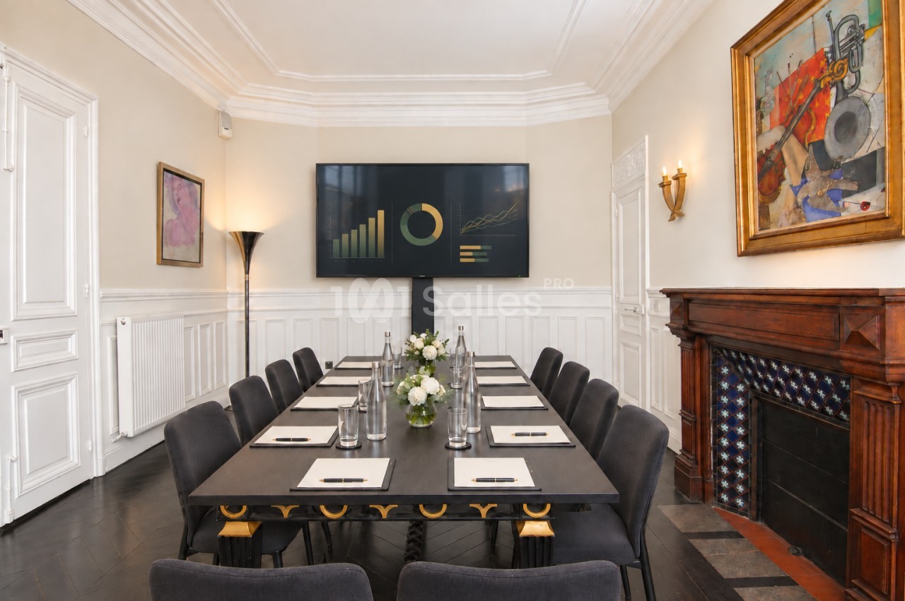 Salle de réunion élégante avec table rectangulaire, chaises noires, écran mural et cheminée décorative.