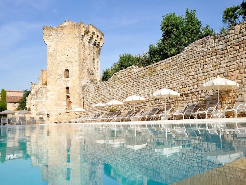 Vue d'une piscine bordée de chaises longues et parasols, avec une tour en pierre ancienne et un mur en arrière-plan.