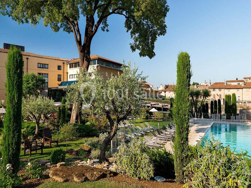 Jardin paysager avec piscine, chaises longues, oliviers et bâtiments en arrière-plan sous un ciel dégagé.