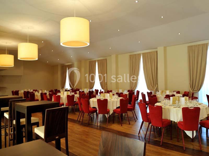 Salle de réception lumineuse avec tables rondes dressées, chaises rouges et grandes fenêtres avec rideaux beiges.
