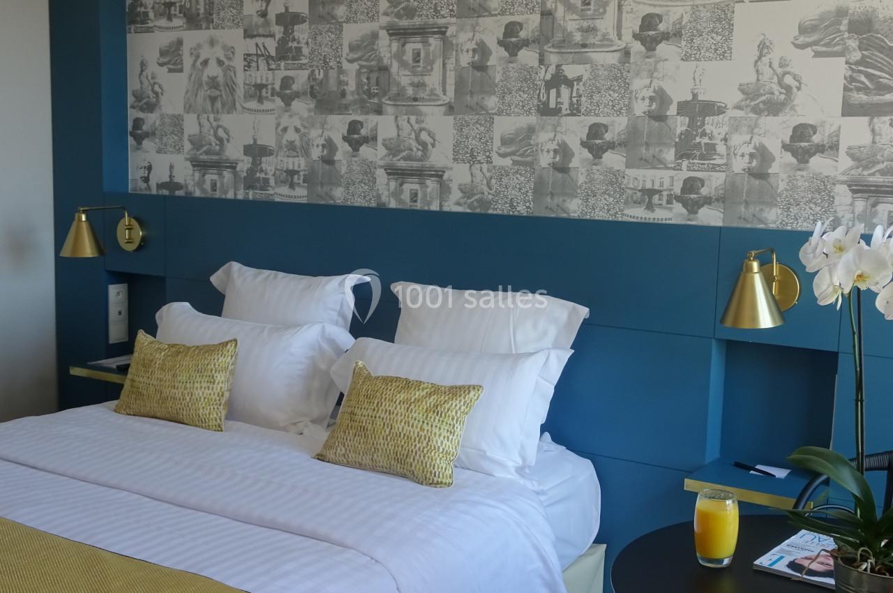 Lit double avec coussins jaunes, mur bleu, papier peint à motifs, lampes dorées et table avec un verre de jus d'orange.