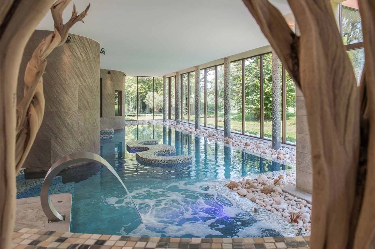 Piscine intérieure avec jets d'eau, entourée de baies vitrées offrant une vue sur un jardin verdoyant.