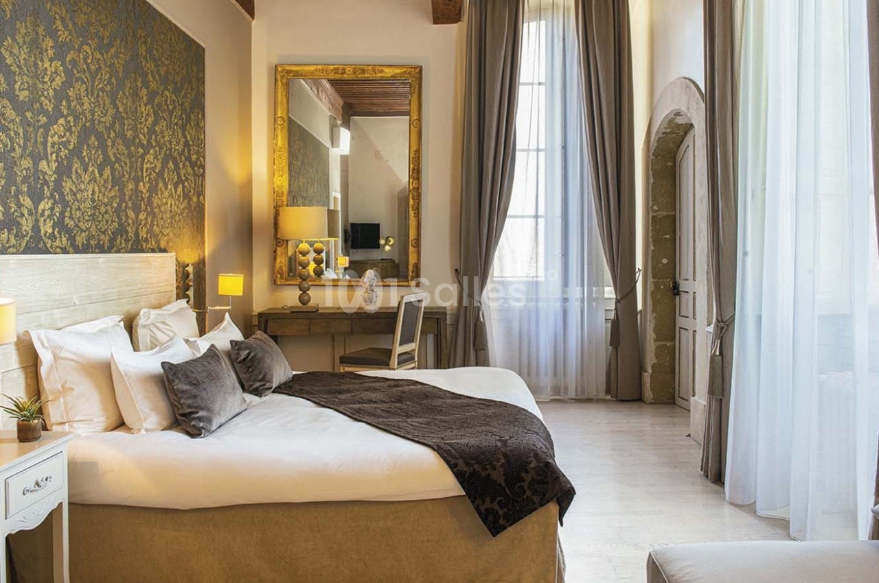 Chambre lumineuse avec lit double, décor élégant, murs ornés de motifs dorés et grande fenêtre avec rideaux.