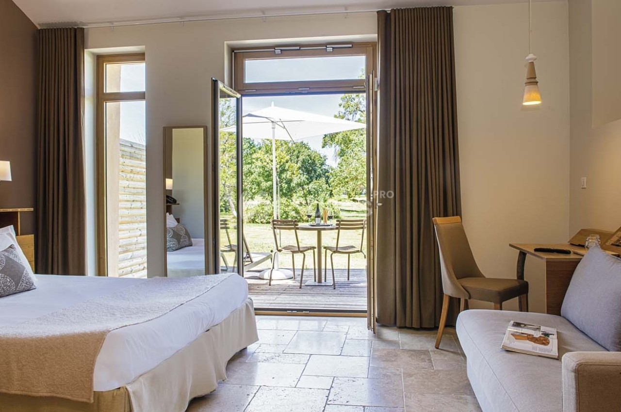 Chambre lumineuse avec lit double, canapé, bureau, et accès à une terrasse donnant sur un jardin verdoyant.