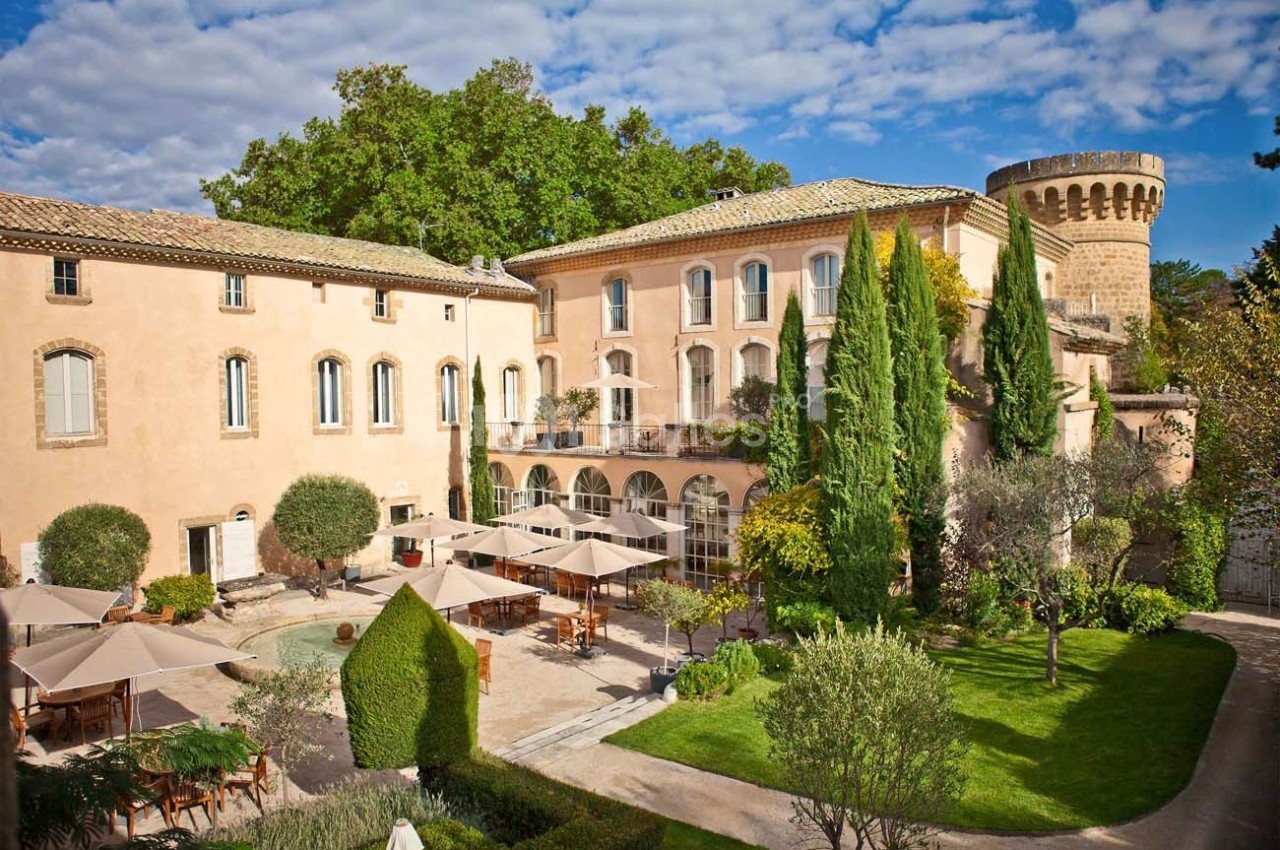 Vue d'un domaine avec bâtiments en pierre, terrasse ombragée, jardin arboré et tour en arrière-plan.