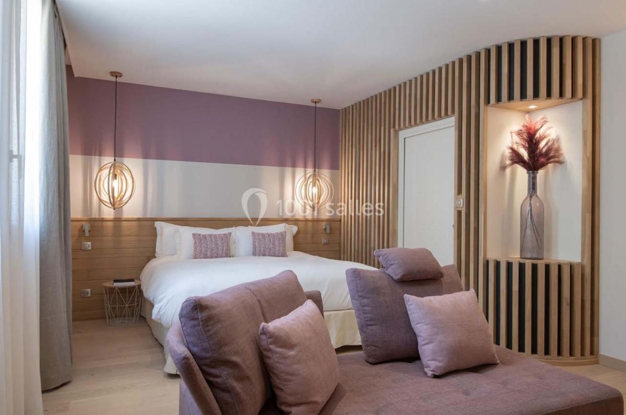 Chambre moderne avec lit double, fauteuils violets, éclairage suspendu et niche décorative avec vase et fleurs séchées.